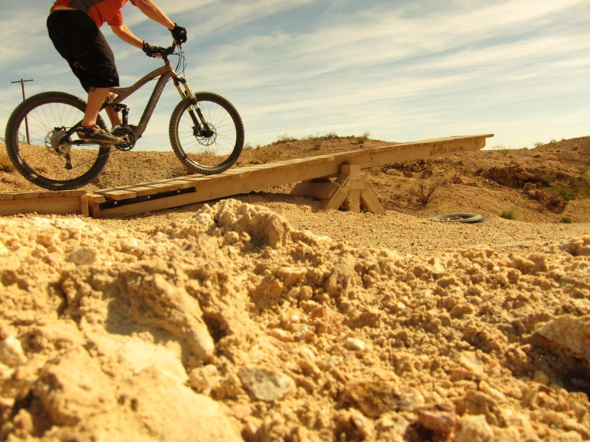 A cyclist riding a mountain bike over a wooden ramp in a desert landscape, with loose sand and gravel in the foreground and a clear blue sky in the background. Bootleg Canyon mountain bike trail.