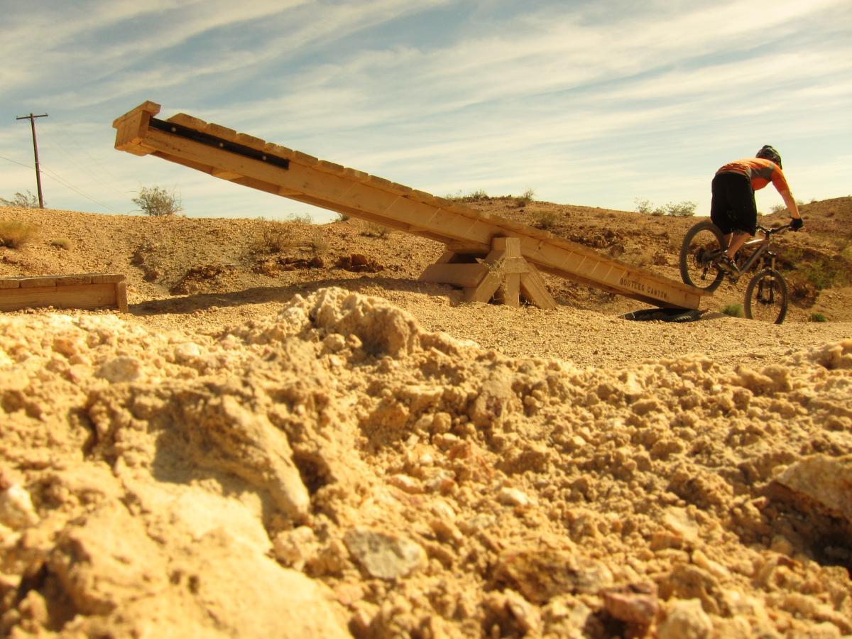 A mountain biker performs a jump off a wooden ramp in a desert landscape, surrounded by rocky terrain and sparse vegetation under a clear blue sky. The foreground shows sandy ground with loose gravel, while the ramp rises at an angle leading to an elevated landing area. Bootleg Canyon mountain bike trail.
