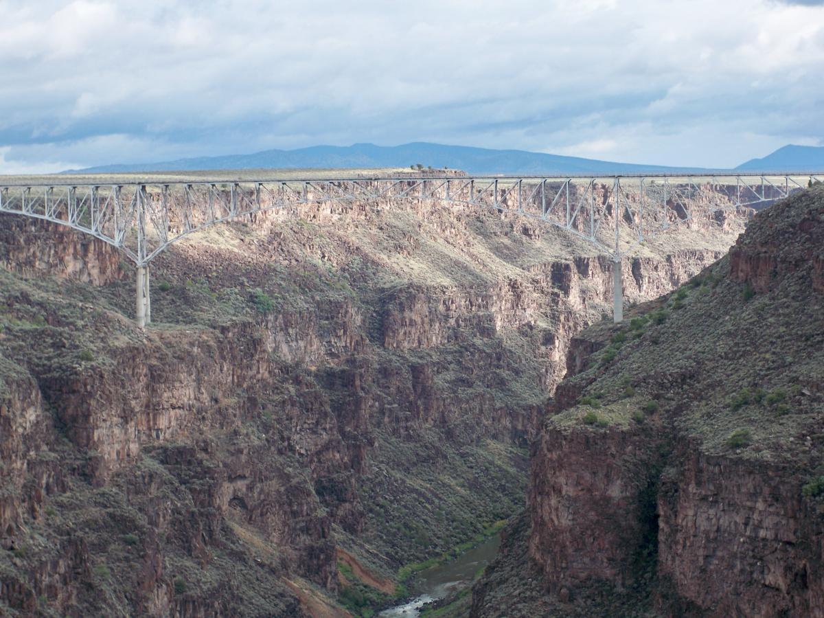 A steel bridge spanning a deep canyon, with rocky cliffs on either side and a river visible at the bottom. The sky is partly cloudy, and the landscape features a mix of greenery and rugged terrain. West Rim, Rio Grande Gorge mountain bike trail.