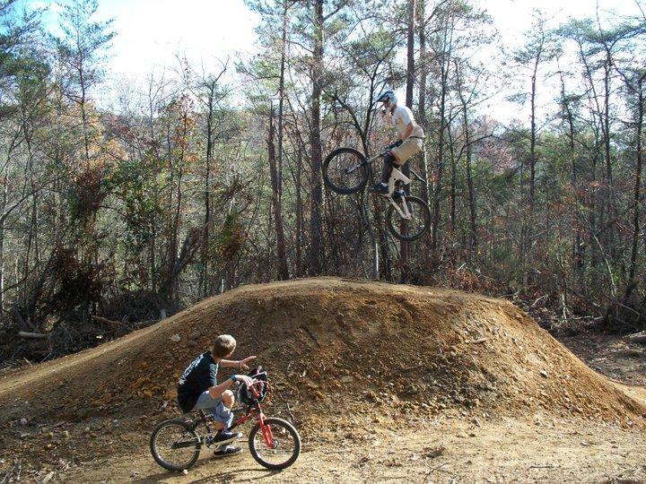 A boy riding a red bicycle observes another cyclist performing a jump off a dirt ramp in a forested area. The jumping cyclist is airborne, showcasing a mid-air trick, while the surroundings are filled with trees and sunlight filtering through the leaves. Hurricane Creek mountain bike trail.