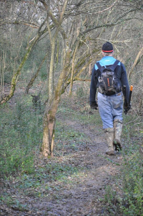 A person wearing a black hat, blue jacket, and rubber boots walks along a dirt path in a wooded area, surrounded by trees and shrubs. They carry a backpack and are heading away from the camera. Lions Club mountain bike trail.
