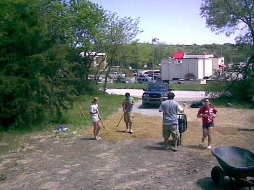 Four individuals engaged in outdoor activity, clearing or organizing a patch of land. They are partially surrounded by greenery and some parked vehicles in the background. One person holds a rake, while others appear to be assisting in various tasks. A wheelbarrow is also visible nearby. The scene is bright, suggesting a sunny day. Warner Park Trails mountain bike trail.