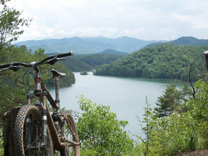 A mountain bike resting on a rocky ledge, overlooking a tranquil lake surrounded by lush green hills and distant mountains under a cloudy sky. Tsali Right Loop mountain bike trail.