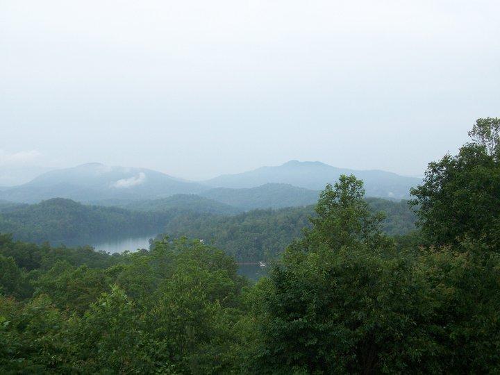 A scenic view of a misty mountain landscape, featuring lush green trees in the foreground, rolling hills in the background, and a serene lake partially obscured by fog. Tsali Right Loop mountain bike trail.