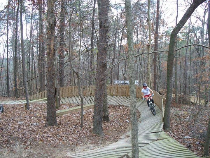 A cyclist navigating a wooden biking trail in a forested area, surrounded by trees with autumn leaves. A sign indicating "Hurricane Creek" is visible in the background. Hurricane Creek mountain bike trail.
