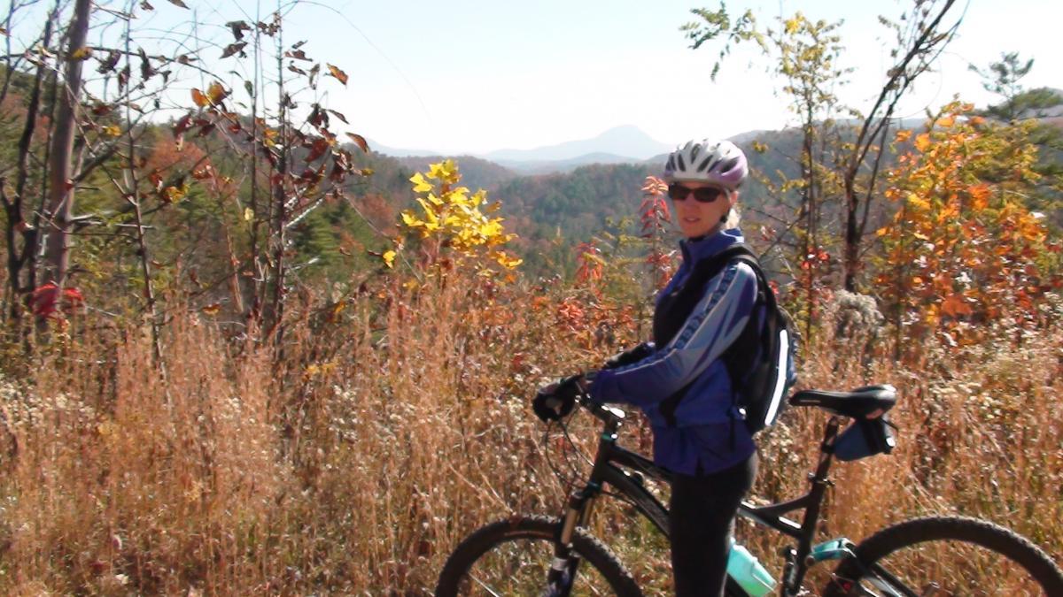 A person in athletic clothing and a helmet stands beside a mountain bike on a trail, surrounded by tall grass and colorful autumn foliage. The background features rolling hills and mountains under a clear sky. Jasus Creek mountain bike trail.