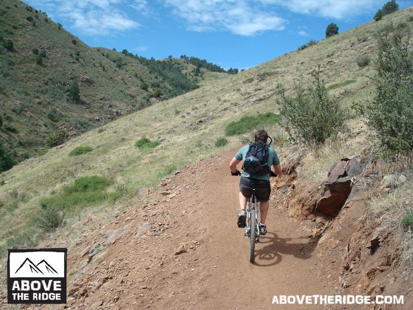 A person riding a mountain bike on a dirt trail surrounded by hilly terrain and green vegetation under a partly cloudy sky. The scene captures the essence of outdoor adventure and cycling in a natural setting. Apex Park mountain bike trail.