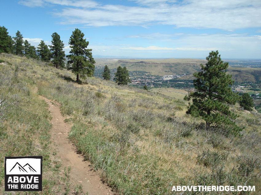 A scenic view of a hiking trail winding through a grassy hillside, lined with tall pine trees. In the background, a valley and distant mountains are visible under a partly cloudy sky. The image features a logo for "Above the Ridge" in the lower left corner. Apex Park mountain bike trail.