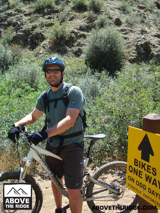 A smiling cyclist wearing a helmet and sunglasses stands beside a mountain bike on a dirt trail. He is dressed in a short-sleeve shirt and shorts, and he is holding the handlebars of the bike. In the background, there is lush vegetation and a sign indicating that bikes are allowed one way on odd days. The sun is shining, suggesting a clear day. Apex Park mountain bike trail.