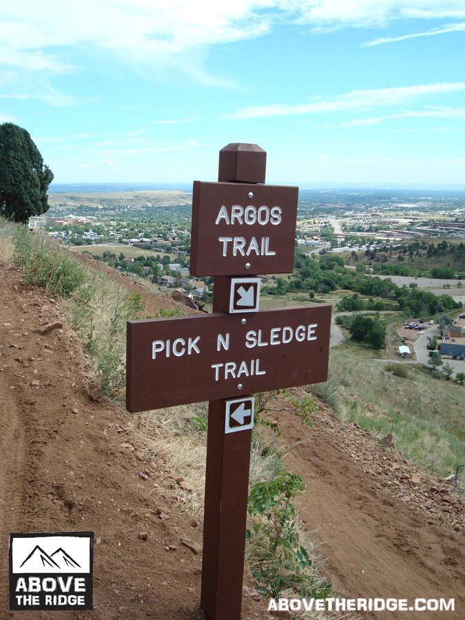 Wooden trail signpost indicating directions for Argos Trail and Pick N Sledge Trail, set against a hilly landscape with a view of a town below and a partly cloudy sky. Apex Park mountain bike trail.