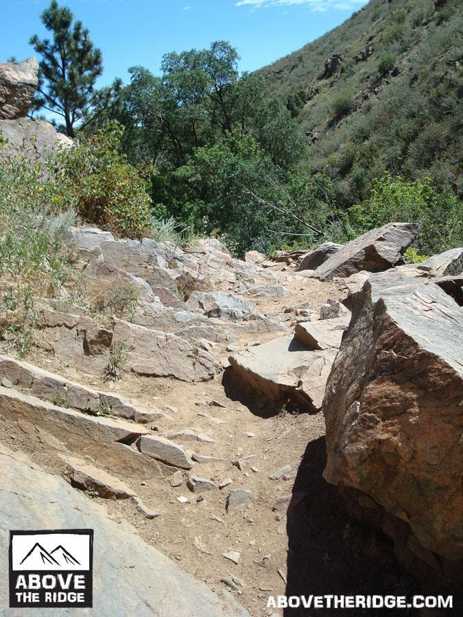 A rocky hiking trail winding through a natural landscape, surrounded by trees and rolling hills under a clear blue sky. The path is lined with uneven stones and dirt, indicating natural wear from foot traffic. Apex Park mountain bike trail.