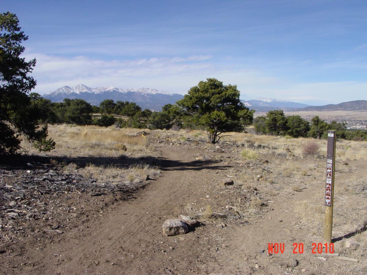 A scenic view of a hiking trail in a grassy area with a few trees, leading towards snow-capped mountains in the distance. A trail marker is visible on the right side of the image, indicating the path. The sky is clear with light clouds, and the landscape features rocky terrain and sparse vegetation. Little Rainbow mountain bike trail.