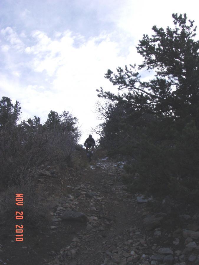 A person riding a mountain bike on a rocky trail, surrounded by sparse vegetation and trees, with a cloudy sky in the background. The image is dated November 20, 2010. Little Rainbow mountain bike trail.
