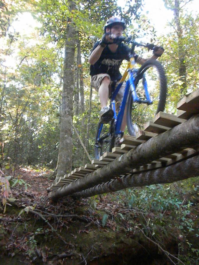 A cyclist mid-jump on a wooden bridge made of logs, crossing a ravine in a wooded area. The bike is blue, and the rider is wearing a helmet and a black t-shirt. Surrounding trees have green leaves with some autumn foliage visible on the ground. Fort Rucker mountain bike trail.