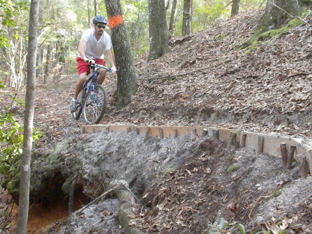 A cyclist wearing a helmet and sunglasses navigates a narrow dirt trail surrounded by trees, leaping off a wooden bridge that spans a small ravine. The forest floor is covered with fallen leaves, and the terrain appears uneven and natural. Fort Rucker mountain bike trail.