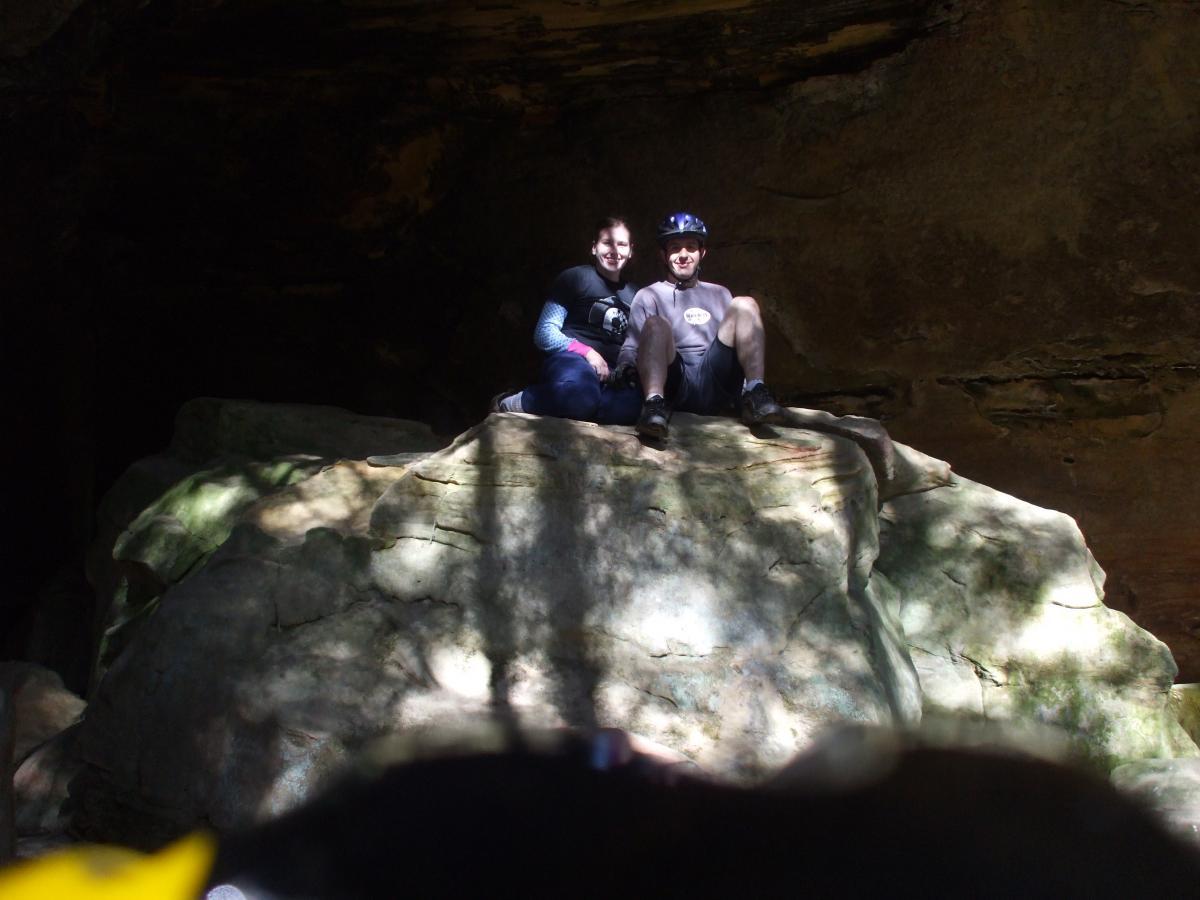 Two people sitting on a large rock inside a cave, with shadows and natural stone formations in the background. One person is wearing a helmet and both are smiling, enjoying their outdoor adventure. Sunlight filters into the cave, illuminating the scene. Laural Lake To Cumberland Falls (sheltowee Trace) mountain bike trail.