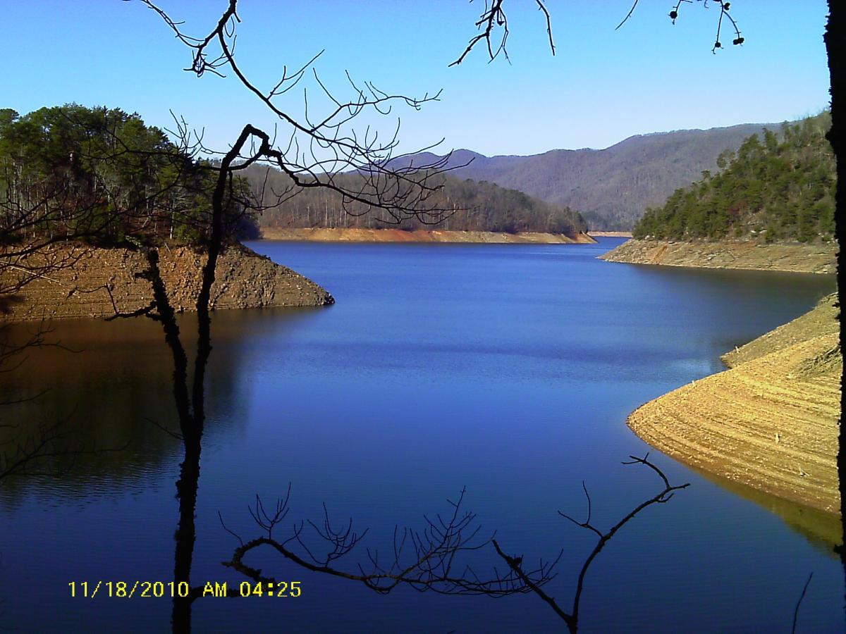A tranquil lake surrounded by hills and trees, with calm blue water reflecting the clear sky. The foreground features branches reaching out over the water, while the distant landscape shows gentle slopes under a bright blue sky. The scene captures a peaceful natural setting. Tsali Recreation Area mountain bike trail.