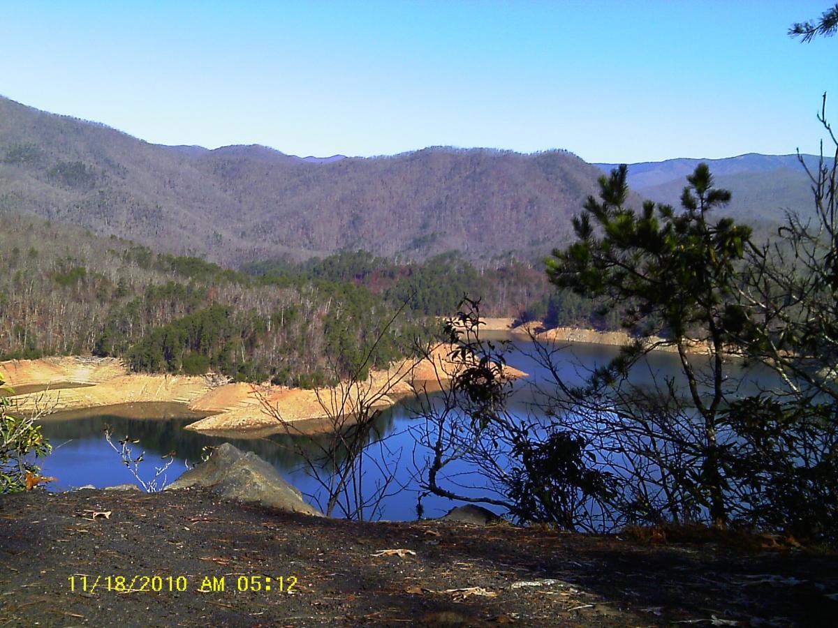 A scenic view of a tranquil lake surrounded by mountains and forests, showcasing calm waters reflecting the landscape under a clear blue sky. In the foreground, rocky terrain and sparse vegetation are visible, while the distant mountains create a picturesque backdrop. Tsali Recreation Area mountain bike trail.