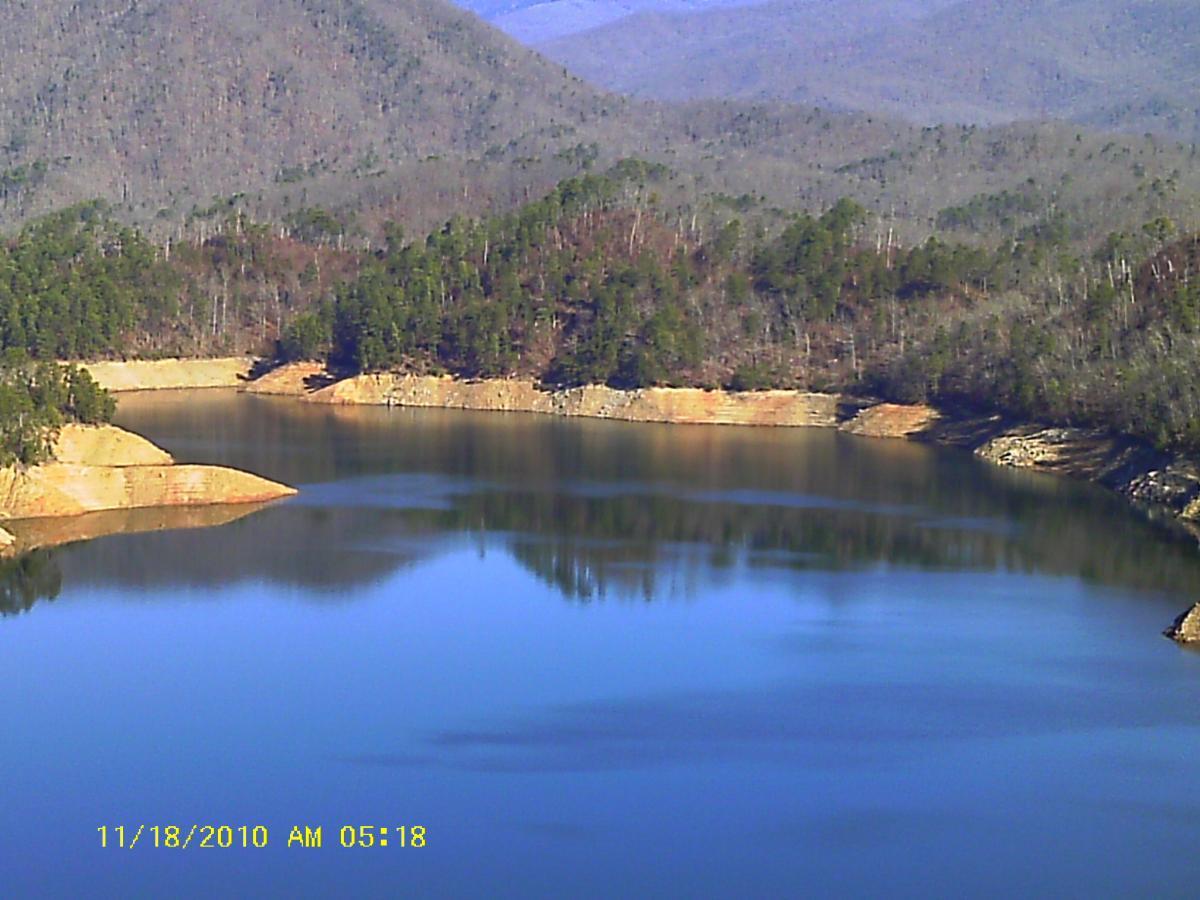 A tranquil lake surrounded by wooded hills and mountains, reflecting the calm water on its surface. The shoreline features sandy and rocky areas, with trees partially covering the hills. The image captures the serene beauty of nature during early morning hours. Tsali Recreation Area mountain bike trail.