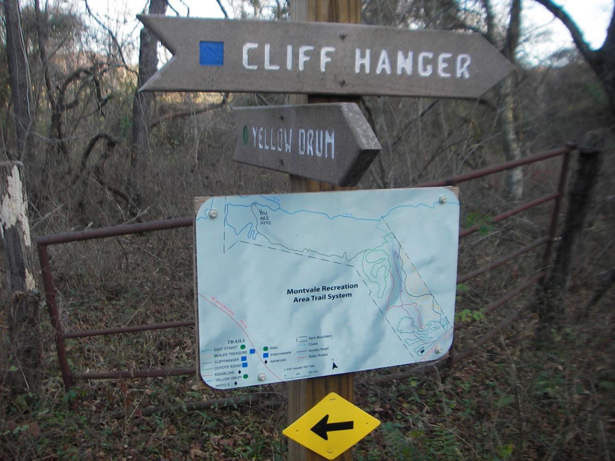 Signpost at the Montvale Recreation Area displaying trail directions, including "Cliff Hanger" and "Yellow Drum," alongside a map indicating the trail system with labeled paths and a marker showing the current location, "You Are Here." Surrounding vegetation and a metal gate are visible in the background. Montvale Trails mountain bike trail.