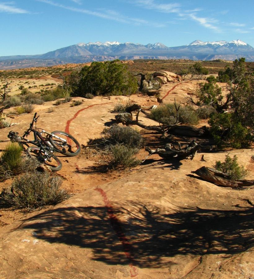 A scenic outdoor landscape featuring a rocky terrain with two mountain bikes leaning against a boulder. In the background, snow-capped mountains rise against a clear blue sky, with sparse vegetation and desert shrubs scattered across the foreground. A distinctive red trail mark runs along the rocky surface. Moab Brand Trails mountain bike trail.