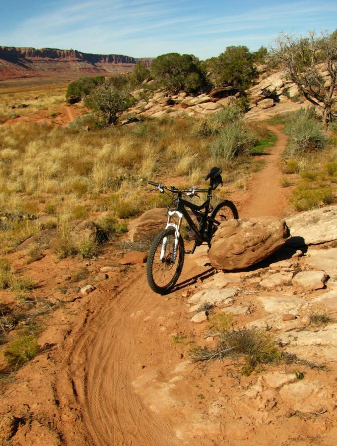 A mountain bike is parked on a dirt trail surrounded by rocky terrain and sparse vegetation, with a backdrop of red rock formations under a clear blue sky. Moab Brand Trails mountain bike trail.