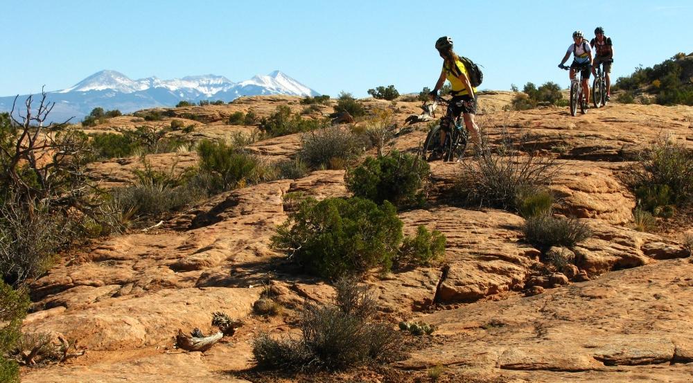 Three mountain bikers ride over rocky terrain with sparse vegetation, set against a backdrop of snow-capped mountains under a clear blue sky. Moab Brand Trails mountain bike trail.