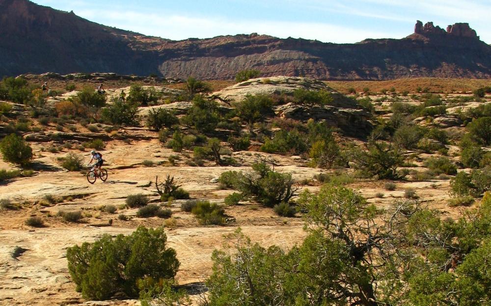 A mountain biker rides along a rocky trail in a vast, arid landscape, surrounded by sparse vegetation and distant rugged hills. The clear blue sky contrasts with the earthy tones of the terrain, showcasing a scenic outdoor environment ideal for biking enthusiasts. Moab Brand Trails mountain bike trail.