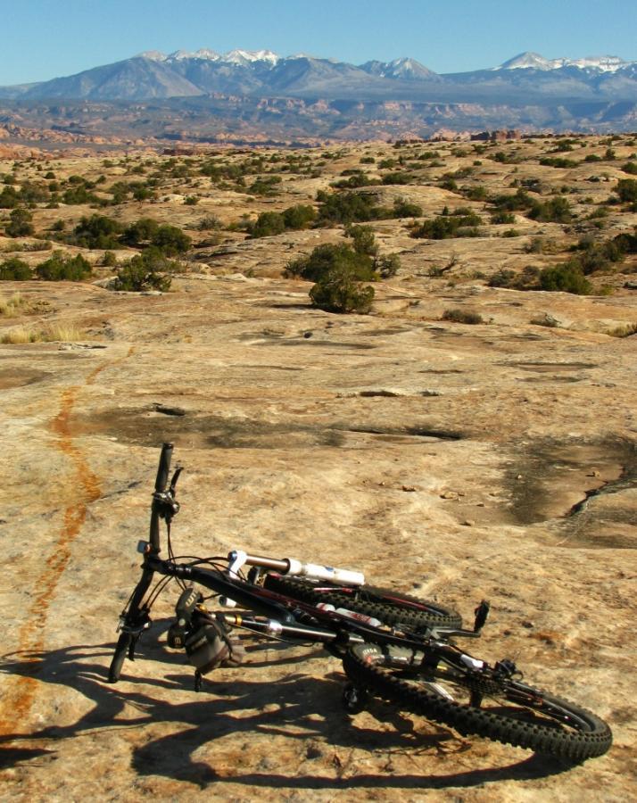 A mountain bike lies on rocky terrain with a backdrop of rugged mountains and blue sky. The landscape features sparse vegetation and scattered boulders, suggesting a remote outdoor area suitable for cycling and adventure activities. Moab Brand Trails mountain bike trail.