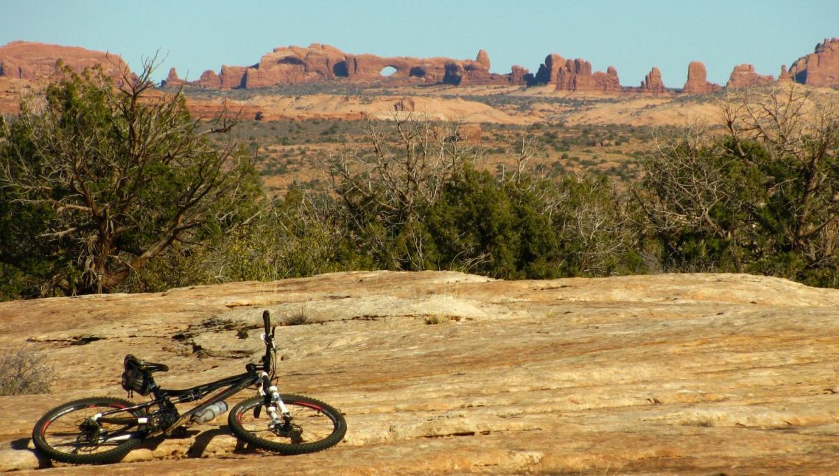 A mountain bike rests on rocky terrain with colorful red rock formations in the background under a clear blue sky. Moab Brand Trails mountain bike trail.