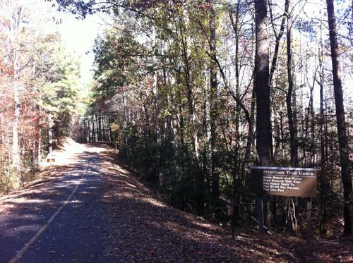 A winding trail surrounded by trees in autumn, featuring colorful foliage and a sign that reads "Attention Trail Users." The pathway is lined with fallen leaves and stretches into the wooded area. Overmountain Victory Trail mountain bike trail.