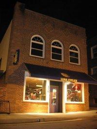 A brick building at night featuring the storefront of a bicycle shop called "Luna Cycles," illuminated by lights, with bicycles visible in the windows and an awning above the entrance.