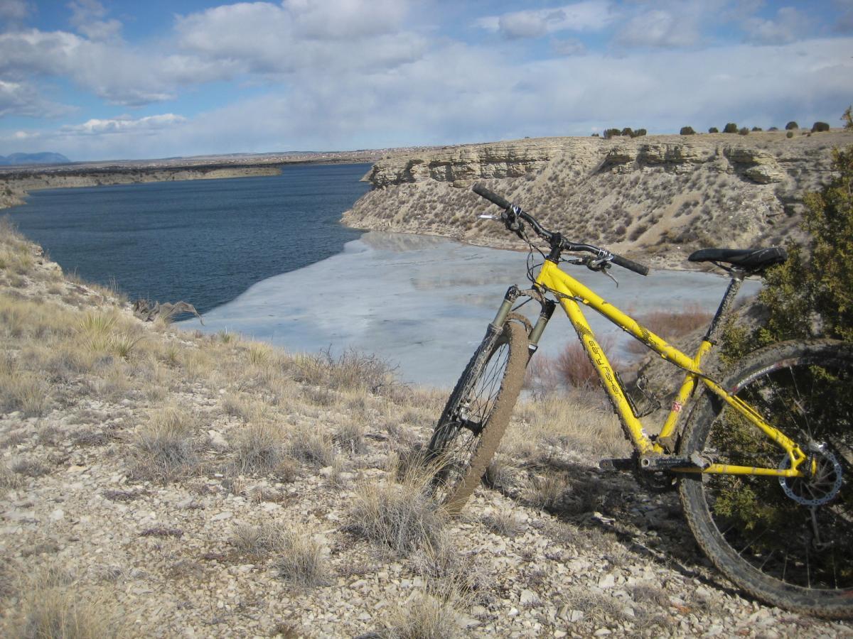 A yellow mountain bike resting on rocky terrain overlooking a calm lake with partially frozen edges. In the background, there are gentle hills and a cloudy blue sky. South Shore Lake Pueblo mountain bike trail.