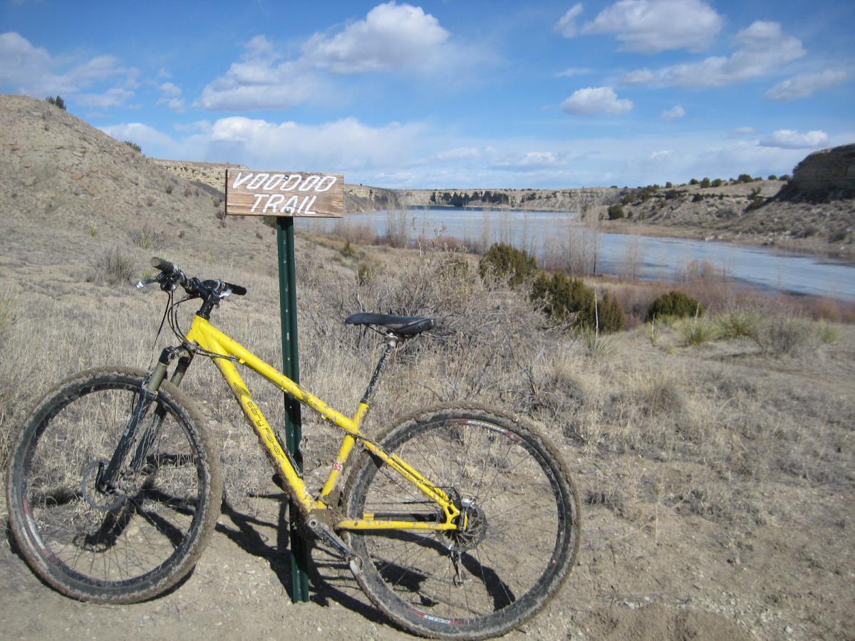 A yellow mountain bike is parked beside a wooden sign that reads "Voodoo Trail." The scene features a rugged landscape with a river in the background, surrounded by dry grass and sparse vegetation under a partly cloudy blue sky. South Shore Lake Pueblo mountain bike trail.
