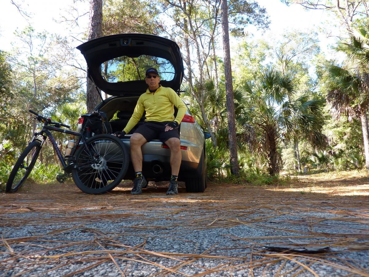A person in a yellow long-sleeve shirt and black shorts sits on the tailgate of a car parked on a gravel trail surrounded by trees and palm plants. A mountain bike leans against the side of the car, and pine needles cover the ground. The scene captures a peaceful outdoor moment, likely before or after a biking adventure. Paisley Woods Trail mountain bike trail.