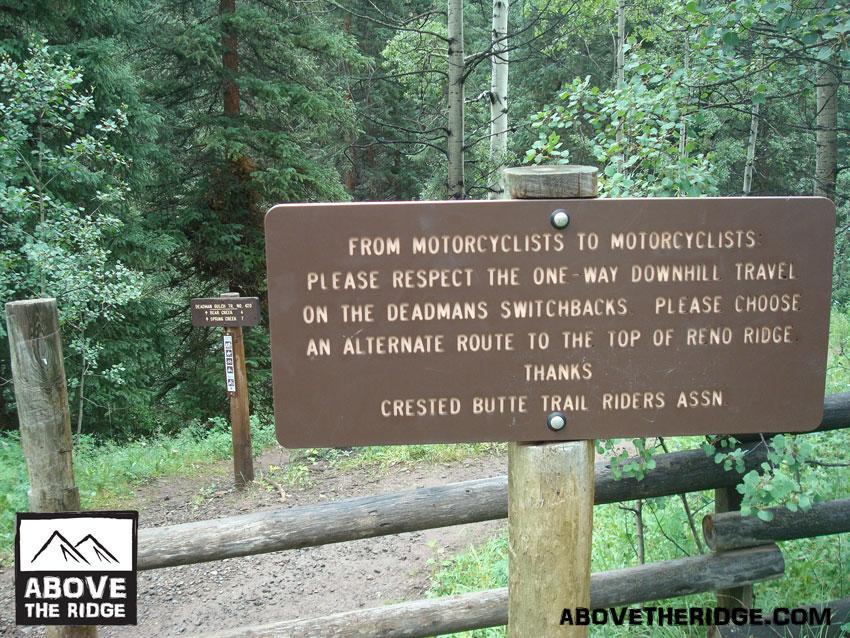 A wooden sign in a forested area provides information to motorcyclists. It advises them to respect the one-way downhill travel on the Deadmans Switchbacks and suggests choosing an alternate route to the top of Reno Ridge. The sign is associated with the Crested Butte Trail Riders Association. The background features lush green trees and foliage. Reno / Flag / Bear / Deadman Loop mountain bike trail.