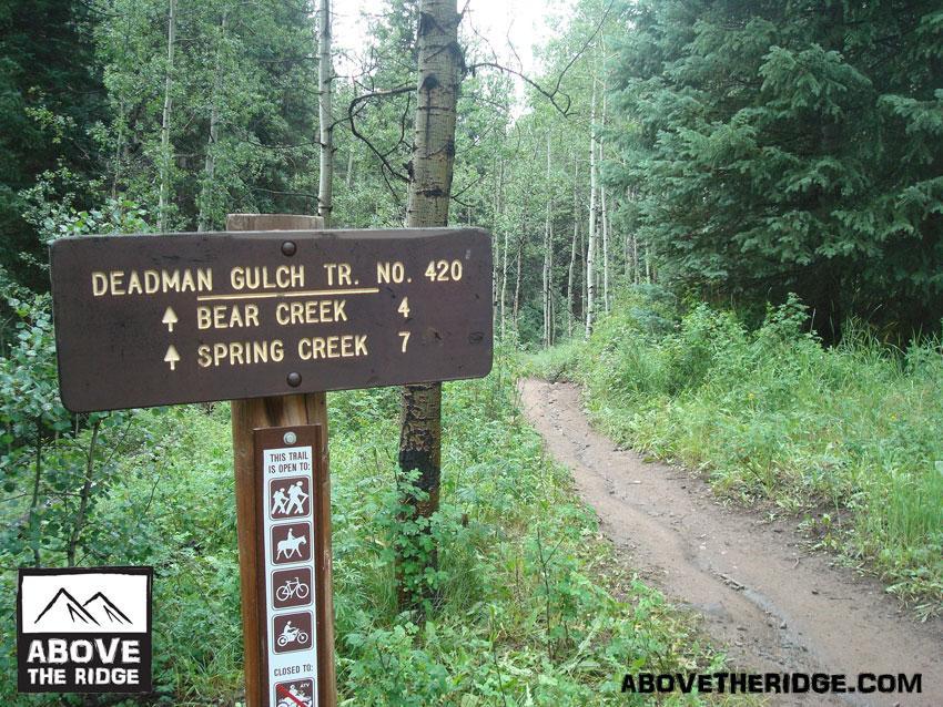 Signpost marking the Deadman Gulch Trail No. 420, indicating distances to Bear Creek (4 miles) and Spring Creek (7 miles), surrounded by a lush forest. The trail is open for hiking, biking, and other outdoor activities. Reno / Flag / Bear / Deadman Loop mountain bike trail.