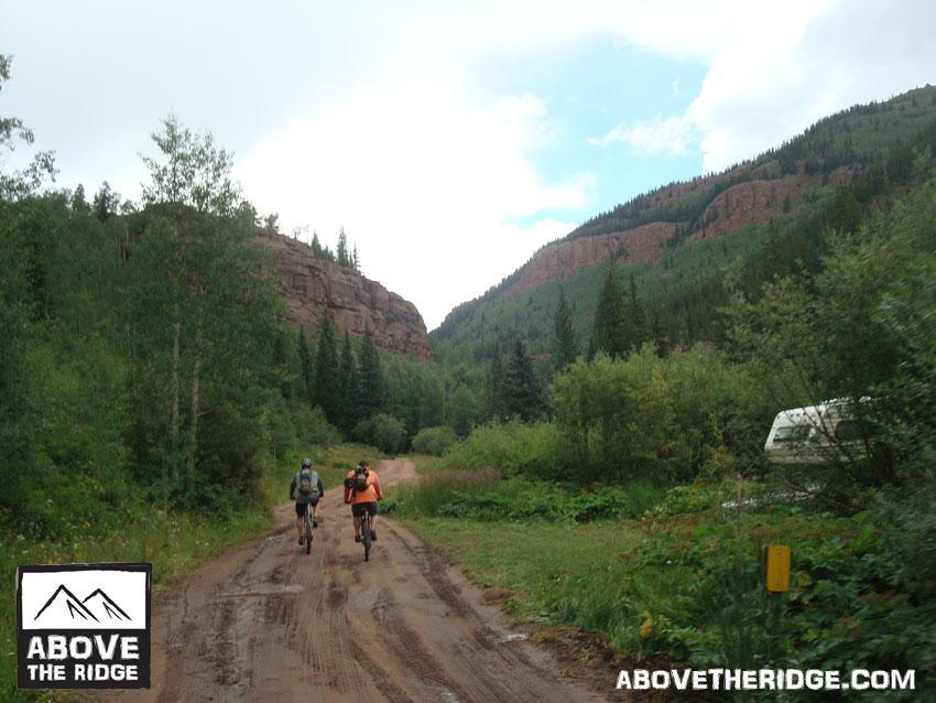 Two mountain bikers riding along a dirt path surrounded by lush greenery and mountains. The sky is partly cloudy, adding a serene atmosphere to the outdoor scene. An RV is visible in the background, indicating a casual recreational setting. The logo for "Above the Ridge" is displayed in the lower left corner. Reno / Flag / Bear / Deadman Loop mountain bike trail.