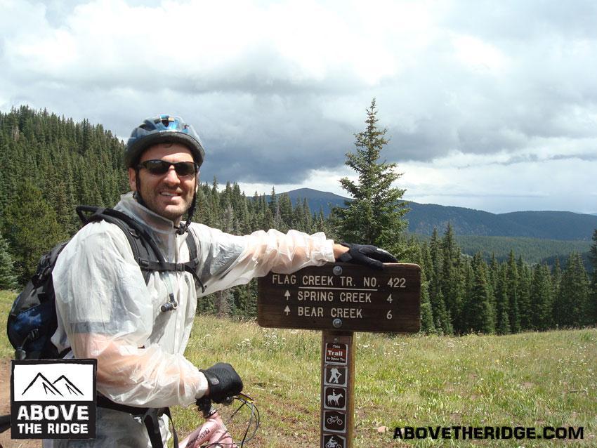 A person wearing a helmet and sunglasses poses next to a wooden trail sign in a mountainous area. The sign shows directions for Flag Creek Trail No. 422, Spring Creek, and Bear Creek, with distances indicated. Lush green trees and a cloudy sky can be seen in the background. The person is dressed in a light rain jacket and is standing next to a bike. Reno / Flag / Bear / Deadman Loop mountain bike trail.