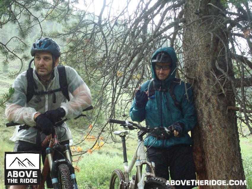Two mountain bikers wearing rain gear stand beside their bikes under a tree during rainy weather. One rider has a serious expression, while the other gestures playfully. The background shows a lush, green landscape with blurred trees. A logo for "Above the Ridge" is displayed in the lower left corner. Reno / Flag / Bear / Deadman Loop mountain bike trail.