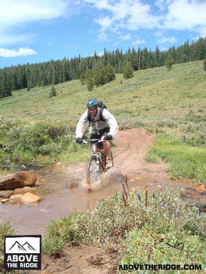 A mountain biker navigates a dirt trail, splashing through a shallow puddle amid a lush green landscape with trees and blue skies in the background. Reno / Flag / Bear / Deadman Loop mountain bike trail.