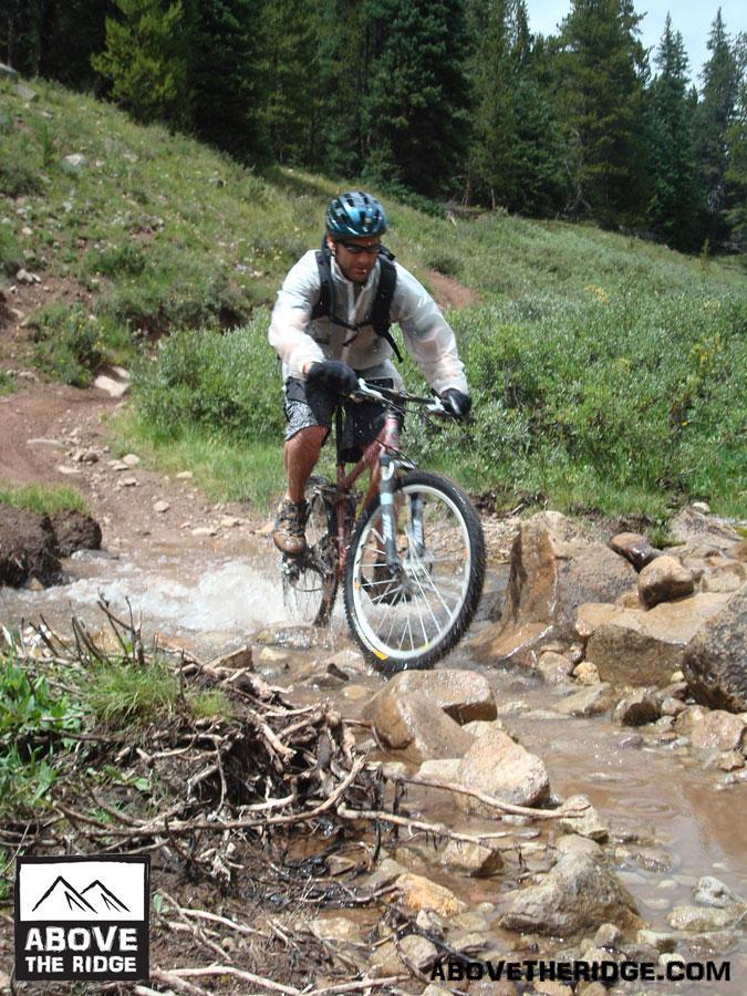 A person riding a mountain bike through a rocky stream in a lush, green forested area. The rider is wearing a helmet and a light rain jacket, navigating over rocks while splashing through water. Reno / Flag / Bear / Deadman Loop mountain bike trail.