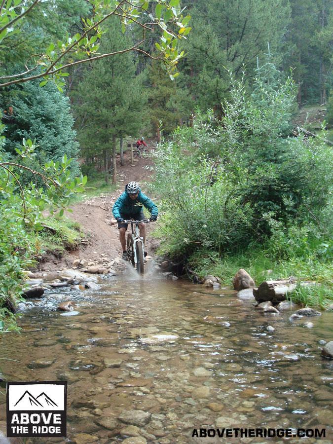 A mountain biker wearing a teal jacket and helmet navigates through a shallow stream on a dirt trail surrounded by lush greenery and trees. Another biker can be seen in the background on the same trail. Reno / Flag / Bear / Deadman Loop mountain bike trail.