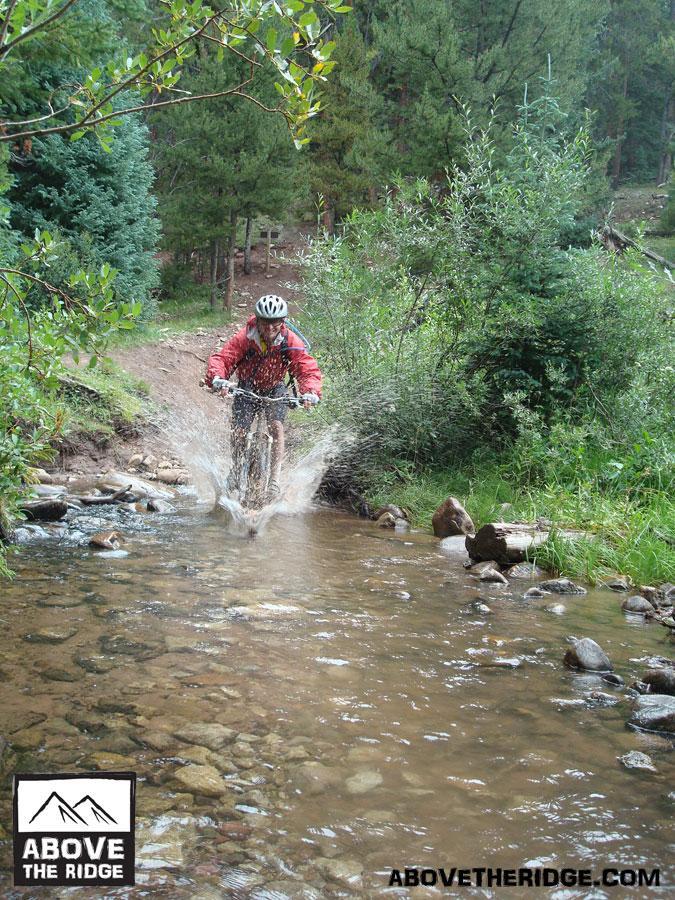 A mountain biker splashes through a shallow creek in a forested trail, surrounded by green foliage and trees. The rider wears a helmet and a red jacket, enjoying the outdoor adventure. Reno / Flag / Bear / Deadman Loop mountain bike trail.