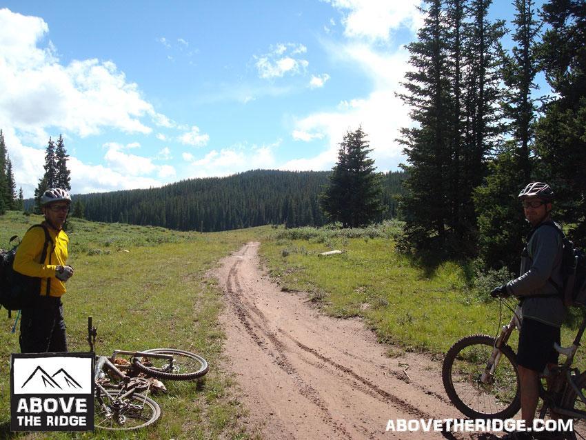 Two mountain bikers stand beside their bikes on a dirt trail in a green, forested landscape under a blue sky with scattered clouds. Pines line the path, which leads into the distance. The scene conveys a sense of adventure and outdoor exploration. Reno / Flag / Bear / Deadman Loop mountain bike trail.