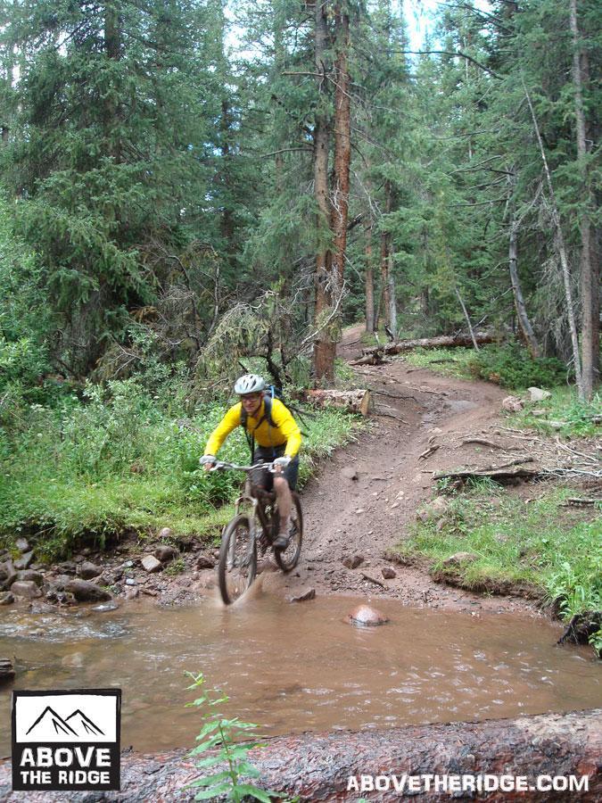 A mountain biker in a yellow jersey navigates a muddy trail through a dense forest, splashing through a small stream. Tall pine trees surround the path, creating a lush, natural setting for outdoor adventure. Reno / Flag / Bear / Deadman Loop mountain bike trail.