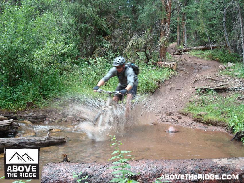 A mountain biker splashes through a shallow creek while navigating a dirt trail surrounded by lush greenery and tall trees. Reno / Flag / Bear / Deadman Loop mountain bike trail.