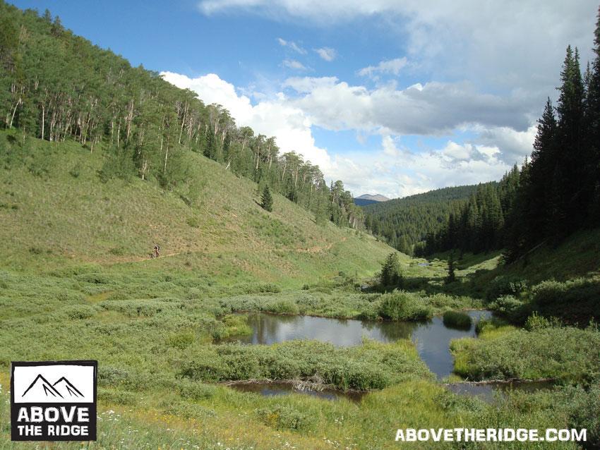 A scenic mountain landscape featuring rolling hills covered in greenery, a natural pond in the foreground, and a hiker on a winding trail. The sky is partly cloudy, with patches of blue peeking through the clouds. Tall trees are visible on the hillsides, creating a serene outdoor atmosphere. Reno / Flag / Bear / Deadman Loop mountain bike trail.