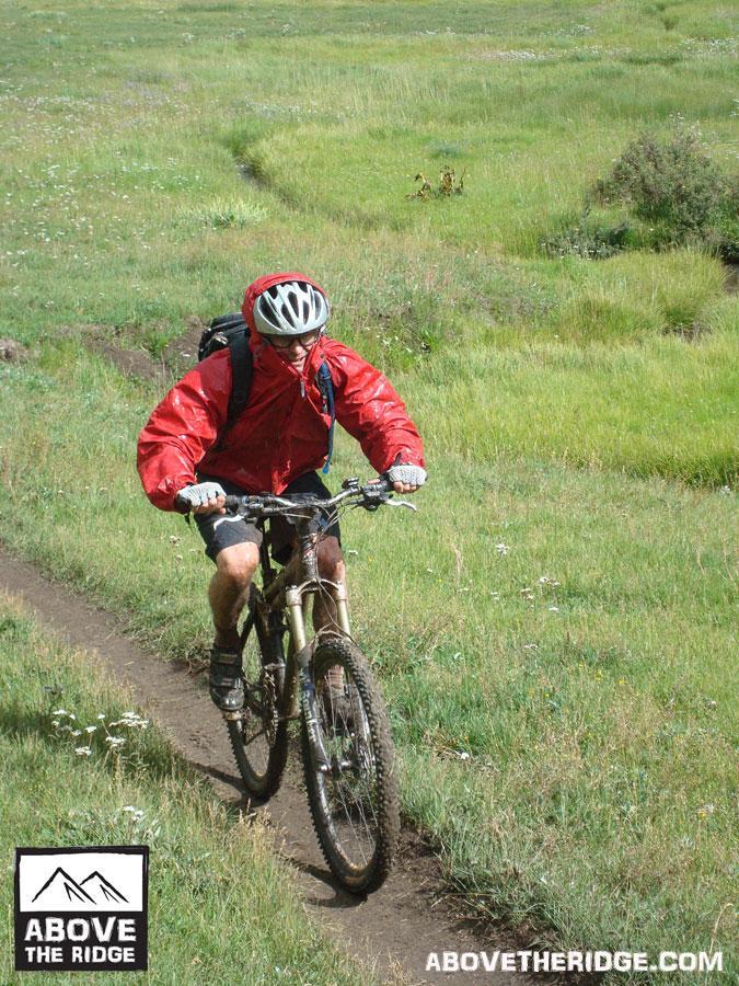 A person riding a mountain bike on a grassy trail, wearing a red rain jacket and a helmet, with a backpack strapped on. The background features a lush green landscape with patches of wildflowers. Reno / Flag / Bear / Deadman Loop mountain bike trail.