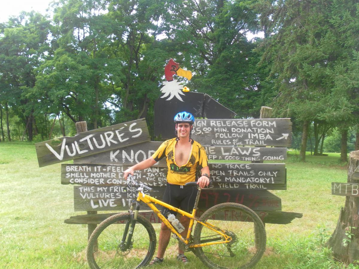 A person wearing a helmet and a yellow t-shirt stands next to a wooden sign that reads "Vulture's Knob." The sign includes trail rules and information about donations. The individual is holding a mountain bike, with trees and grassy terrain in the background, suggesting an outdoor biking area. Vultures Knob mountain bike trail.